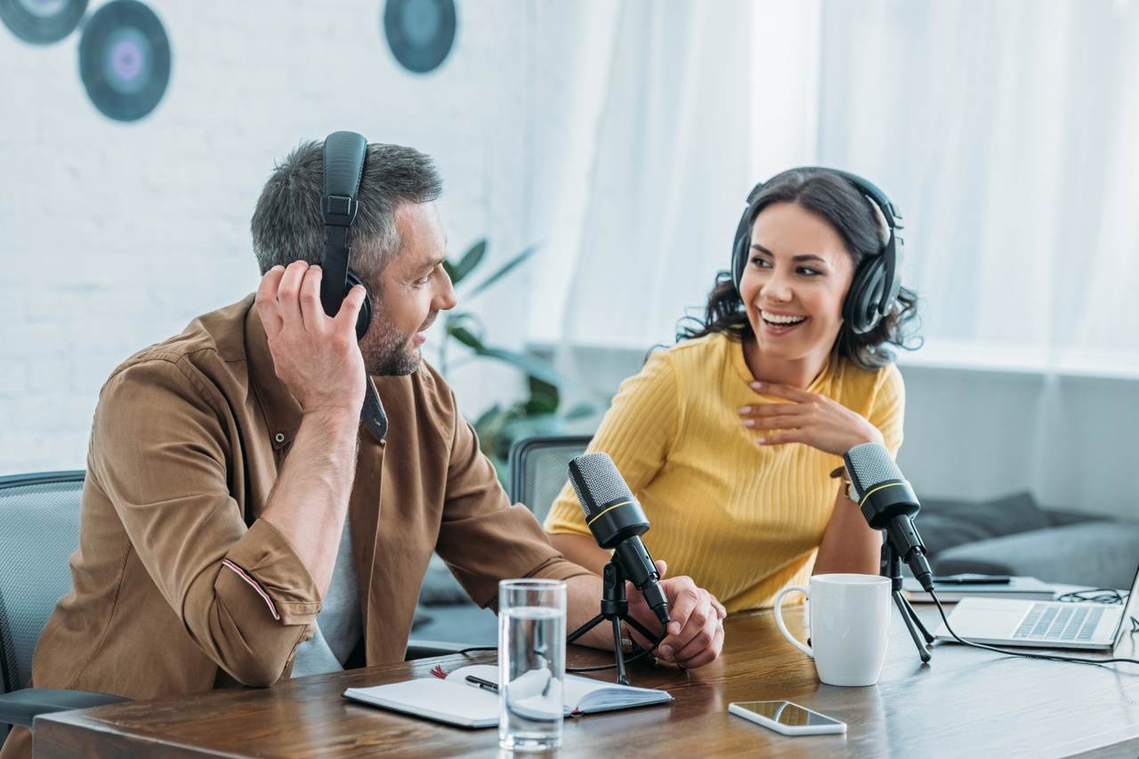 a picture of a man and a woman smilling and looking at each other with headphones on sitting behind a desk and podcasting