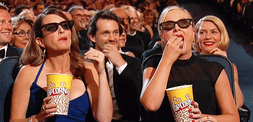 Promi-GIF. Amy Poehler und Tina Fey bei den Emmys sitzen im Publikum mit 3D-Brille und einem Becher Popcorn. Sie mampfen und starren auf die Bühne, vertieft in die Live-Action-Szene, die sie sich ansehen.