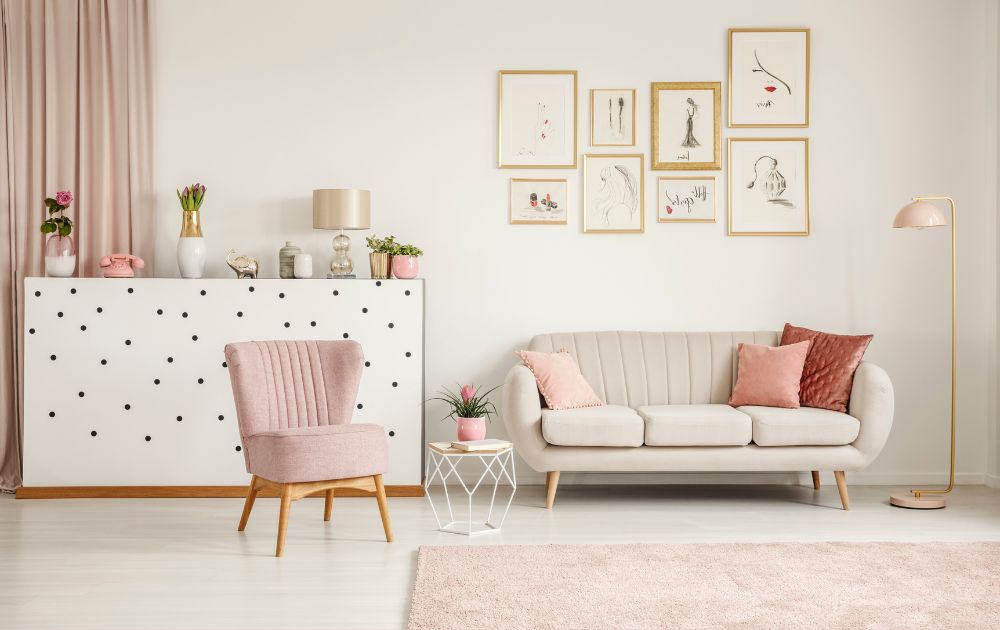 Sala de estar com paredes brancas, cortinas rosa claro, sofá quase branco com almofadas rosa, poltrona rosa claro, console branco com pontos pretos, luminária de pé e desenhos emoldurados em ouro