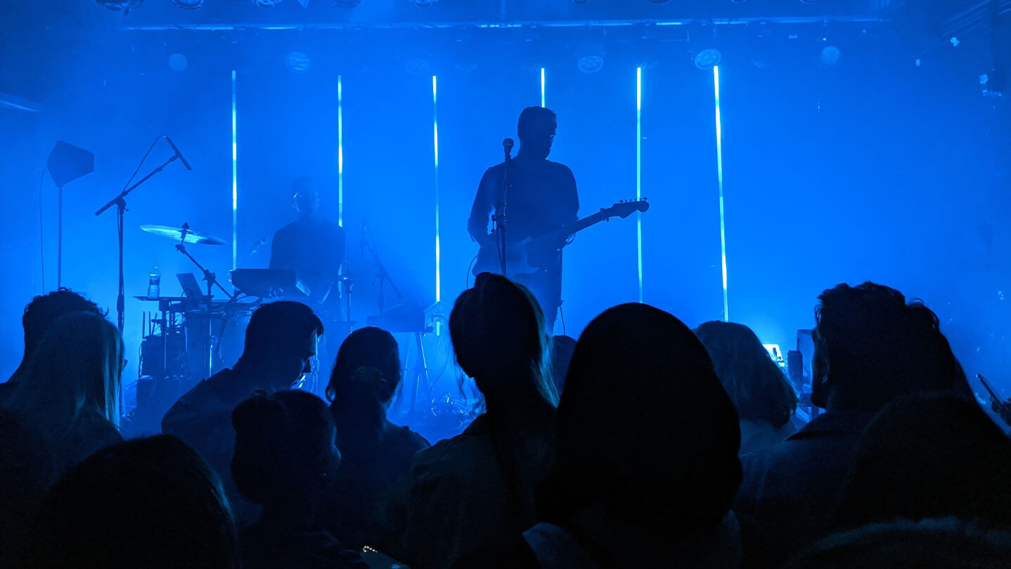 Foto scattata in mezzo a una folla di persone, guardando un chitarrista e un batterista sul palco. Illuminazione scenica blu.