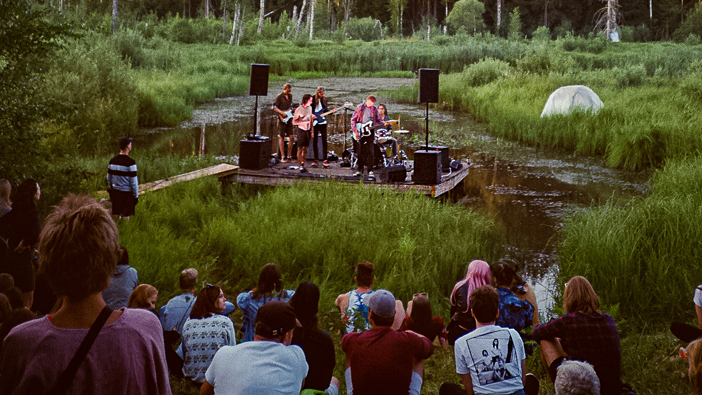 Banda de 6 piezas tocando en un escenario al aire libre construido en un pantano: una pequeña multitud se sienta en la orilla, viendo el espectáculo.