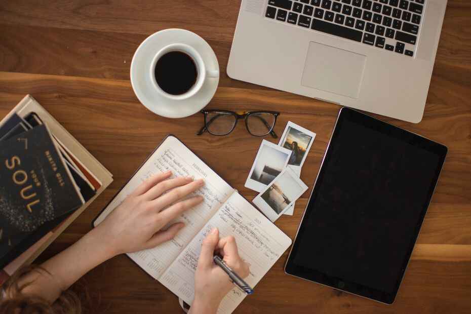 Une personne écrivant dans un cahier, entourée de livres, d'un ordinateur portable, de Polaroids et d'une tasse de café