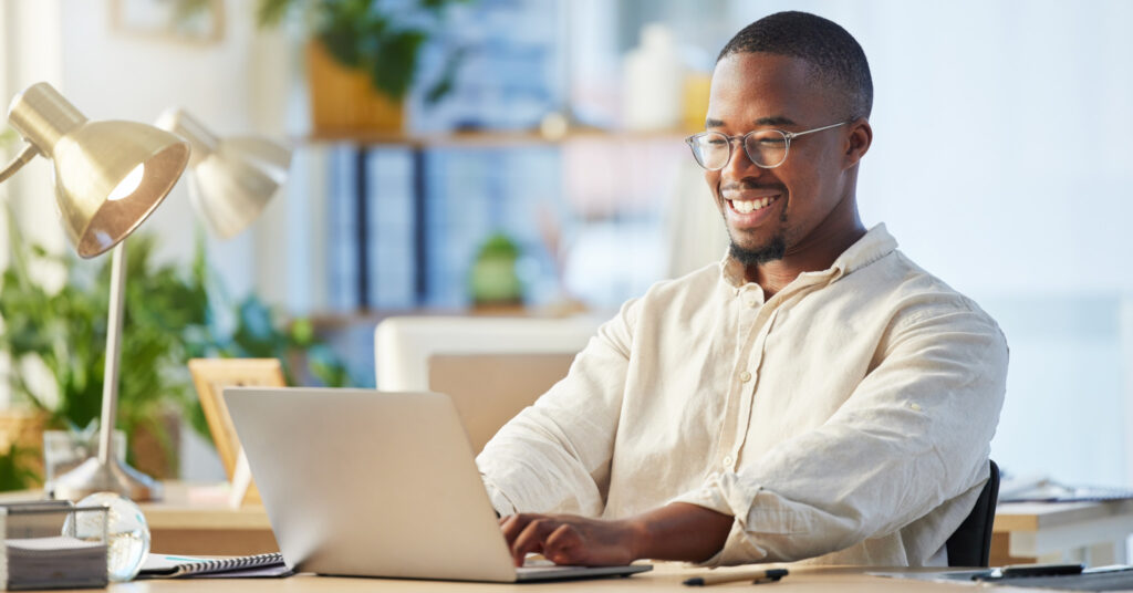 Un profesional negro en una oficina escribe en una computadora portátil mientras sonríe ante el excelente asunto del correo electrónico que está escribiendo.