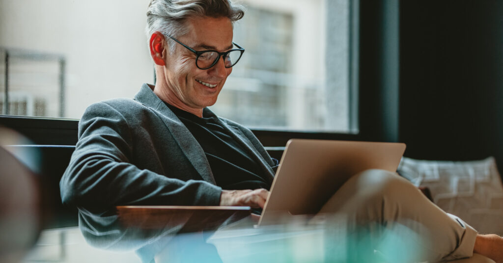 Lächelnder Geschäftsmann sitzt in der Lobby seines Büros und liest tolle E-Mail-Betreffzeilen auf einem Laptop.