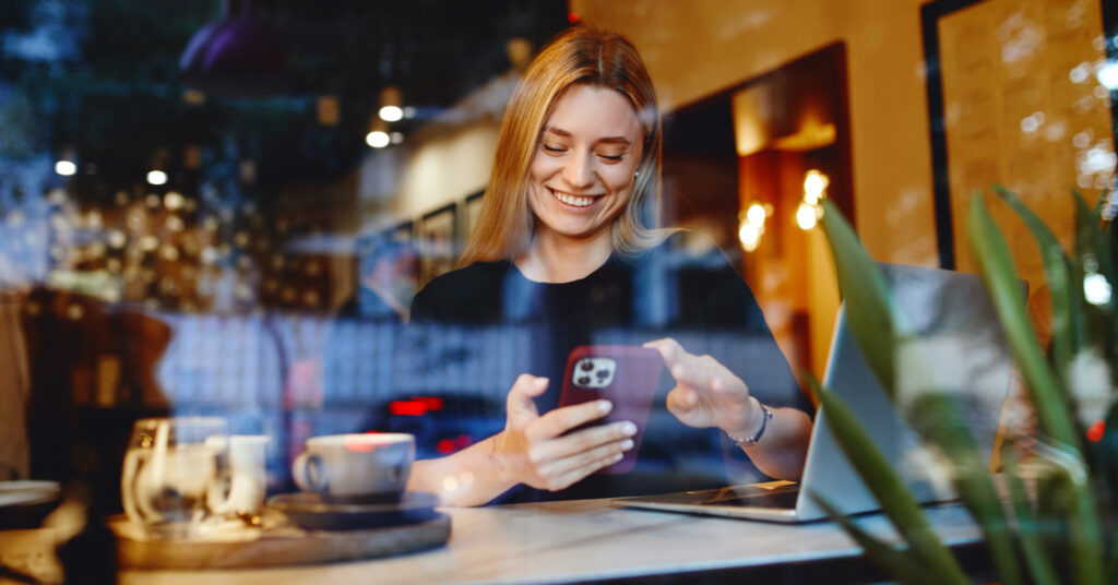 Mujer rubia sentada en una cafetería usando un teléfono inteligente para leer las líneas de asunto del correo electrónico.