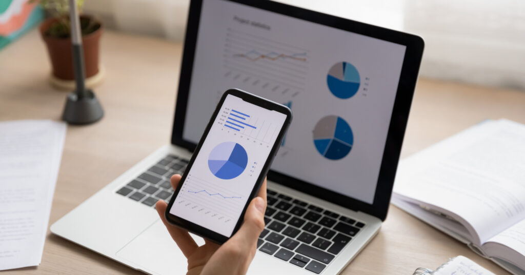 Zbliżenie białej dłoni trzymającej smartfon przed laptopem, oba ekrany przedstawiają grafikę danych