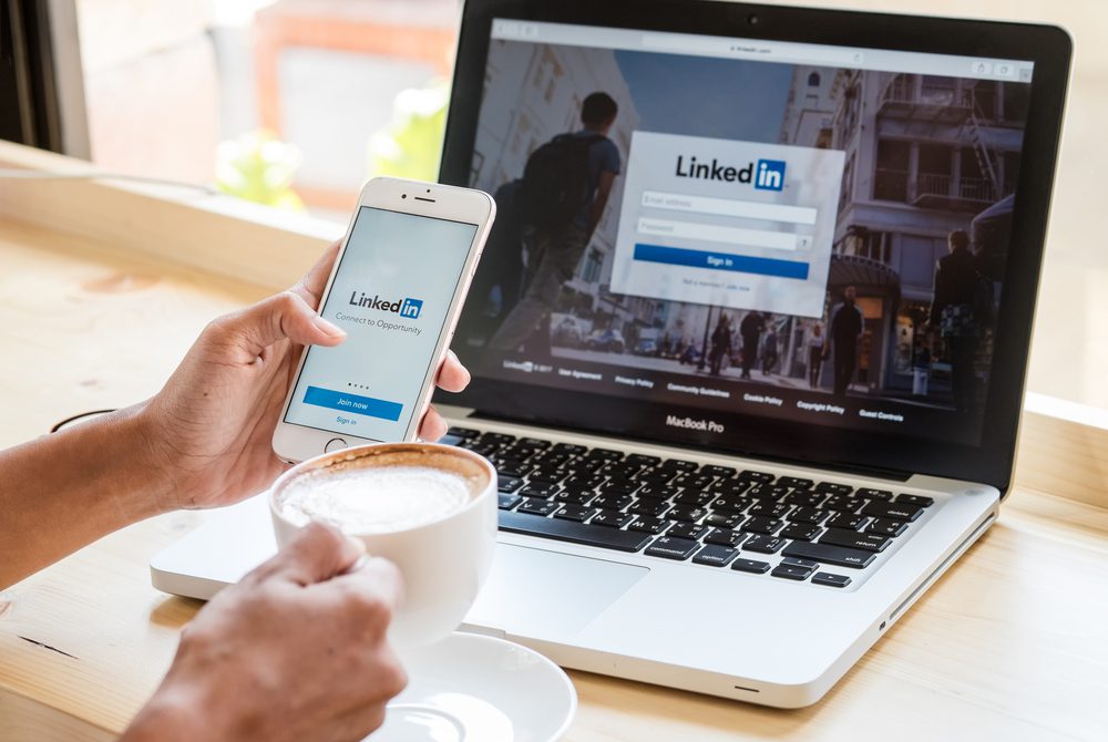 Um profissional de marketing segura um smartphone e um café na frente de um laptop (por algum motivo). Ambos os dispositivos exibem páginas de login do LinkedIn.