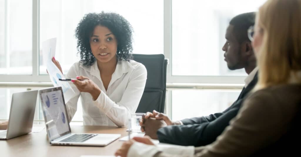 une femme de couleur discute de l'automatisation du marketing des services financiers lors d'une table de conférence ensoleillée avec deux collègues