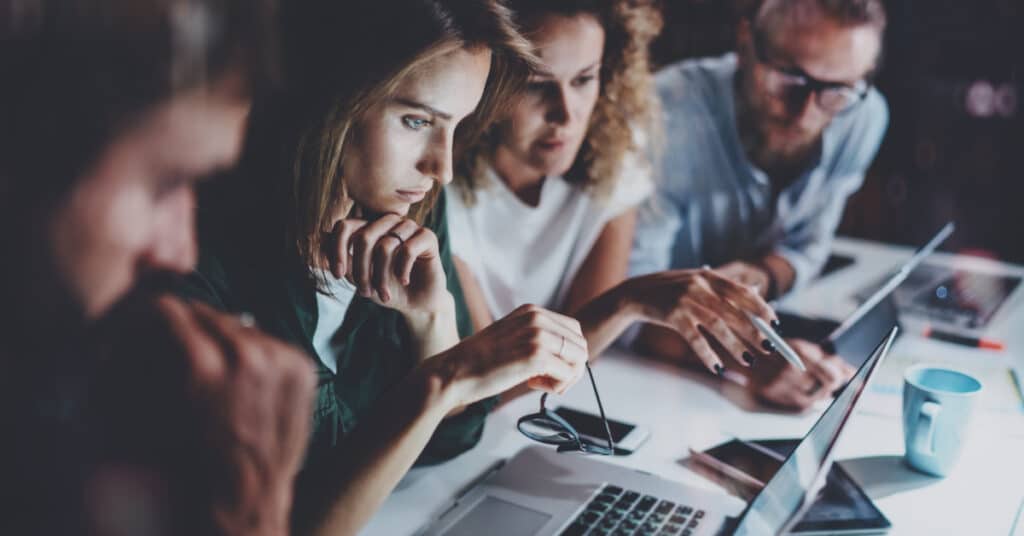 Pemasar yang sibuk berkumpul di depan laptop mereka di kantor yang gelap, menggambarkan struktur departemen pemasaran b2b.