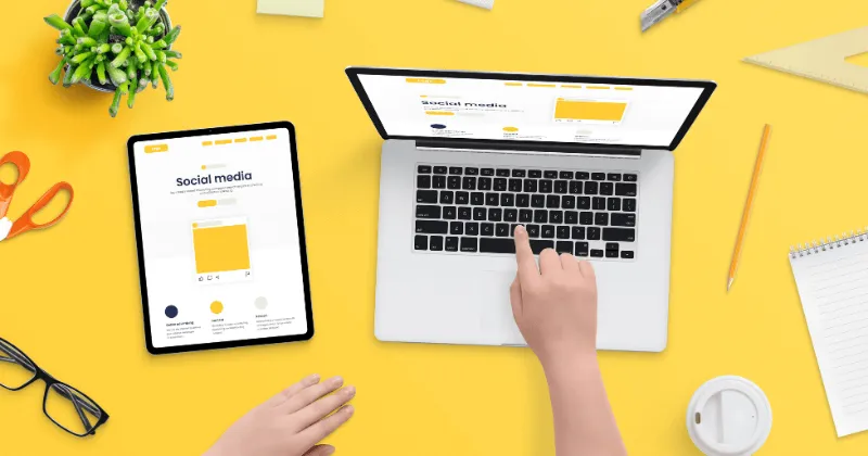 Personne tapant sur un ordinateur portable, une tablette et des fournitures de bureau posées sur un bureau jaune.