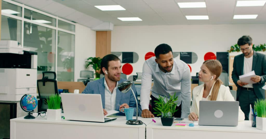 Un collègue se penche pour aider deux autres personnes dans un bureau ouvert, illustrant l'idée des meilleures pratiques d'aide à la vente.