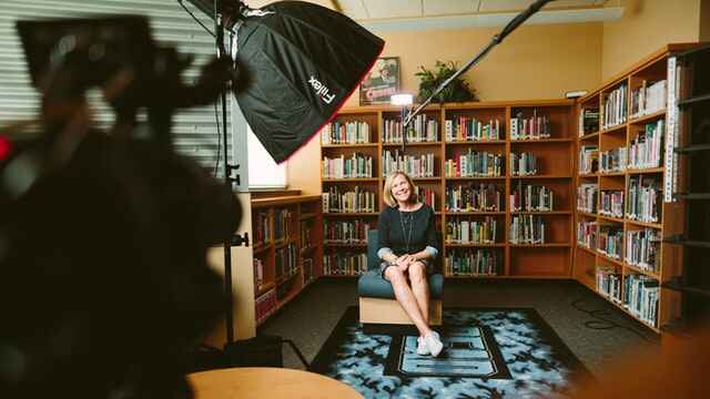 Une femme sourit dans un fauteuil devant une bibliothèque. Devant elle, on peut voir des microphones, un plafonnier et une caméra utilisée pour filmer une interview.