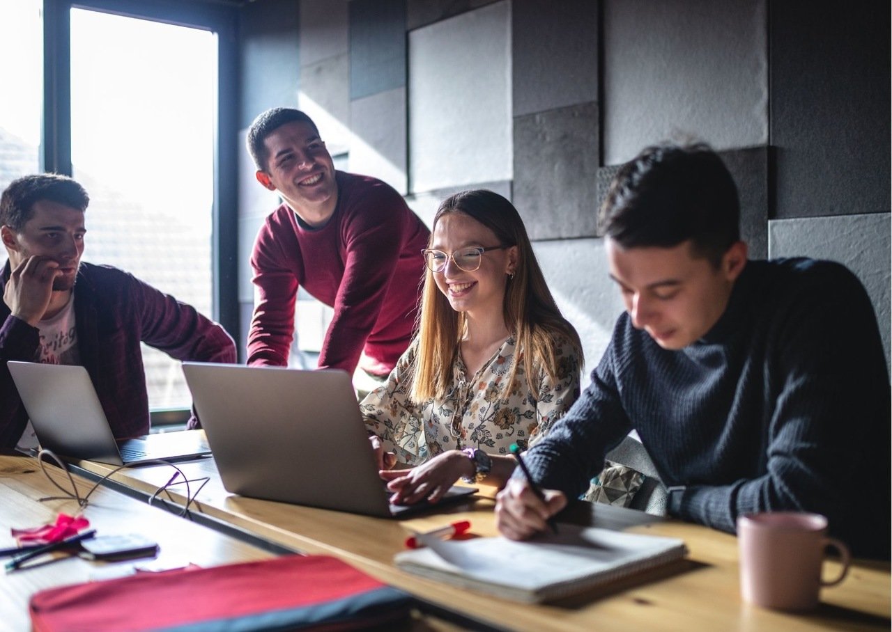 cuatro personas alrededor de la mesa revisando la computadora y riéndose con sinceridad, una está tomando notas mientras la otra da consejos