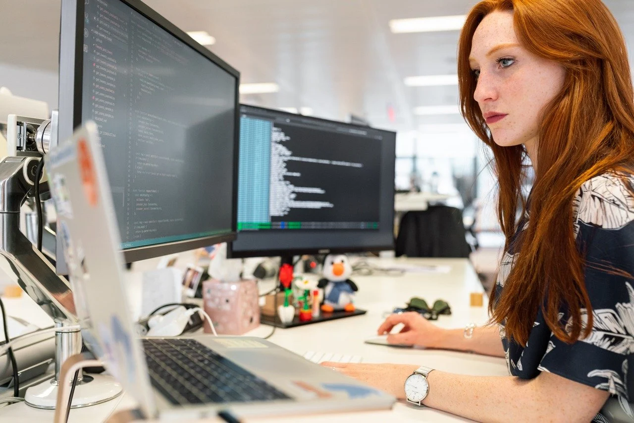 une femme rousse écrivant des codes avec deux moniteurs différents et un MacBook